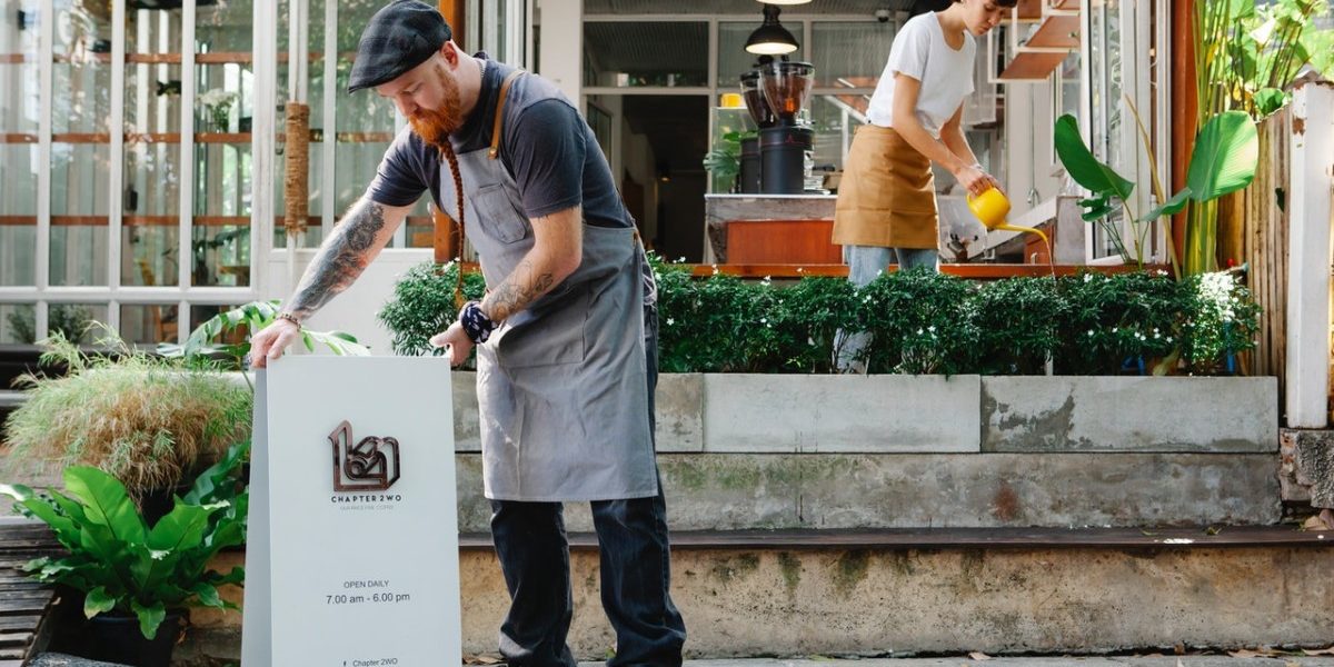 Homme barbu roux avec un béret et un tablier qui installe le panneau signalétique de son restaurant