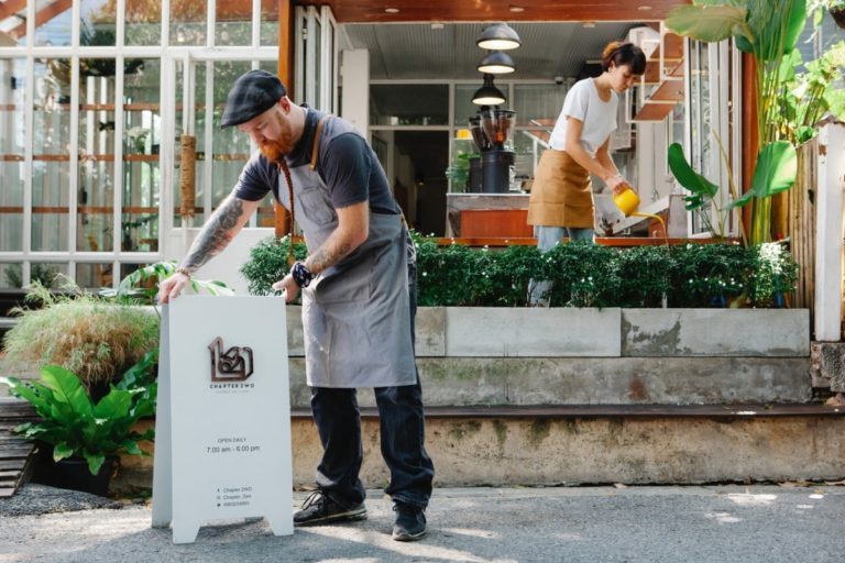 Homme barbu roux avec un béret et un tablier qui installe le panneau signalétique de son restaurant